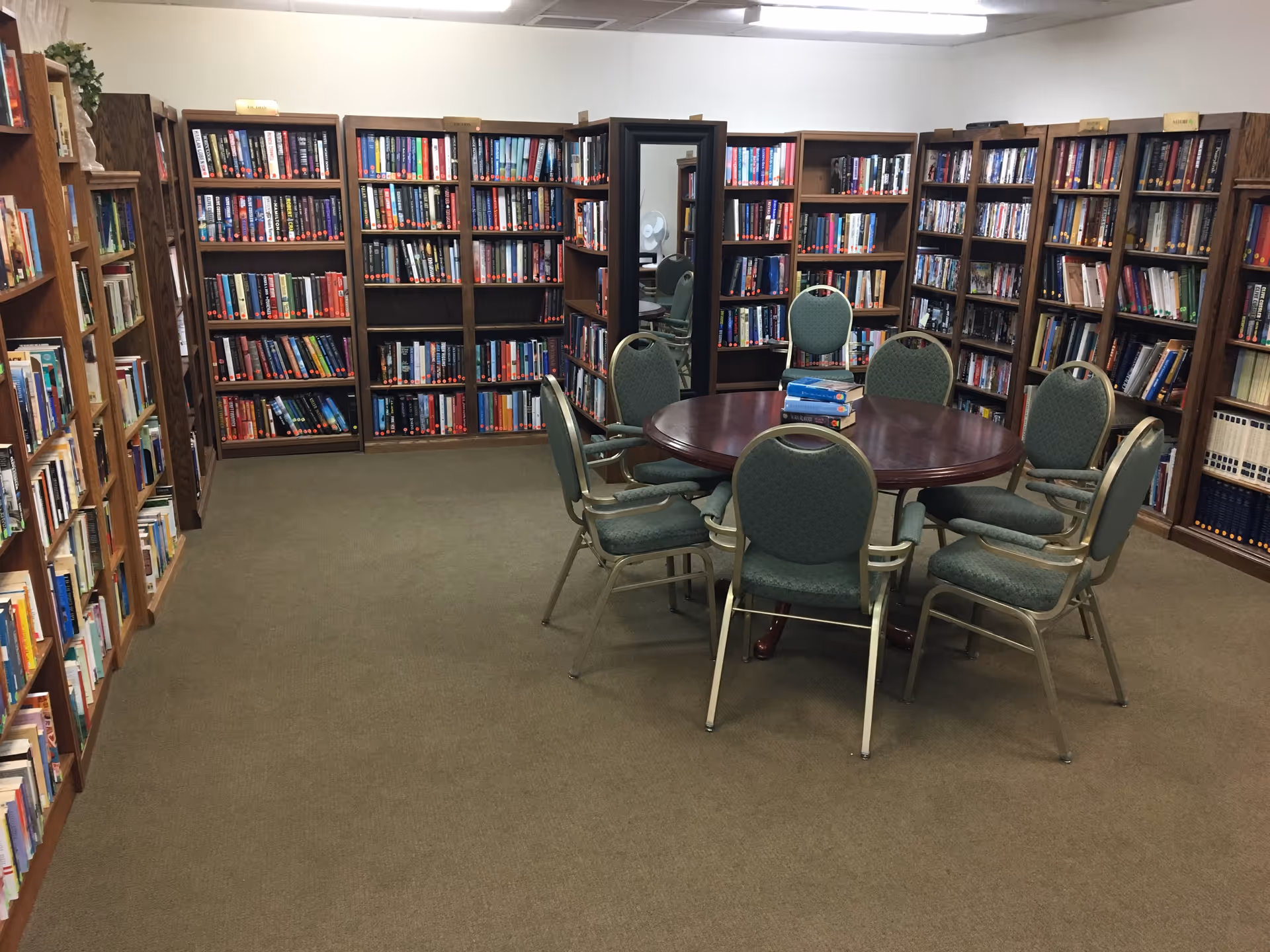 A quiet library room with multiple wooden bookshelves filled with books lining the walls. In the center, there is a round wooden table surrounded by six green cushioned chairs. A few books are stacked on the table. A large mirror is positioned against one bookshelf, reflecting part of the room.