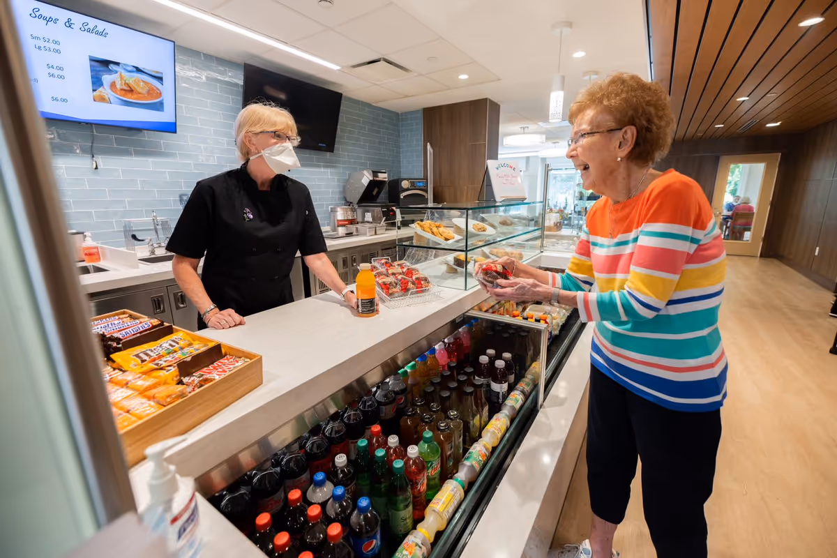 A masked staff member behind a cafe counter serves an elderly woman holding snacks in a senior living community, with refrigerated drinks and baked goods on display.
