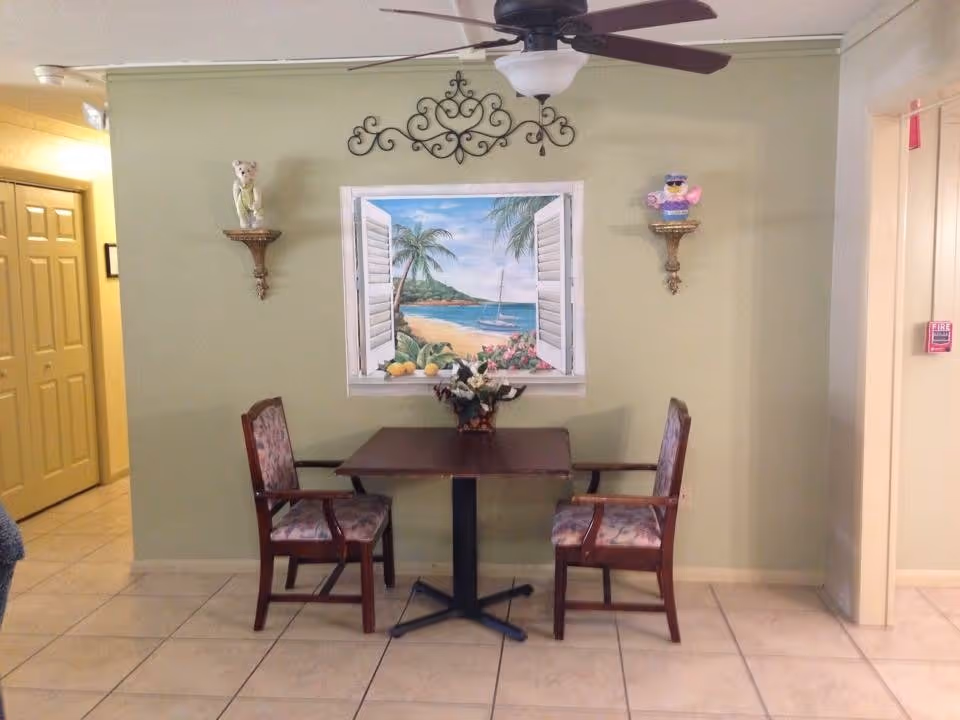 Small seating area with a two-person table and chairs against a green wall decorated with a beach painting, shelves, and a ceiling fan.
