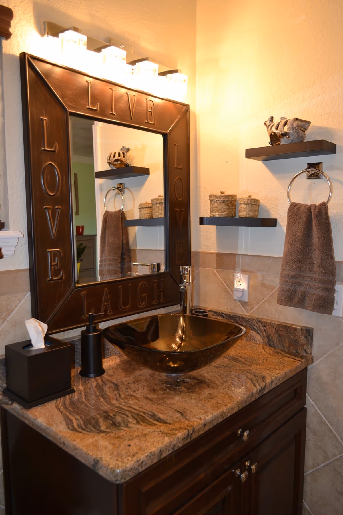 A bathroom vanity with a brown marble countertop, a dark brown vessel sink, and a modern faucet. Above the sink is a large mirror framed with the words LIVE, LOVE, and LAUGH. On the countertop are a black soap dispenser and a black tissue box. To the right, there are two floating shelves with decorative items and a brown hand towel hanging on a chrome towel ring. The walls are tiled halfway up with beige tiles and painted light beige above.