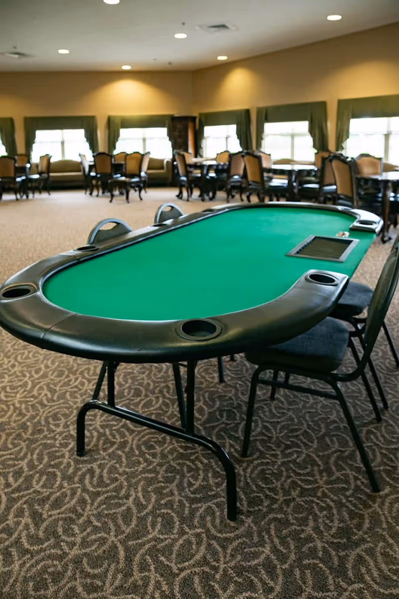 A spacious room with a green felt poker table in the foreground and several dining tables with chairs in the background. The room has patterned carpet, large windows with green curtains, and soft overhead lighting.