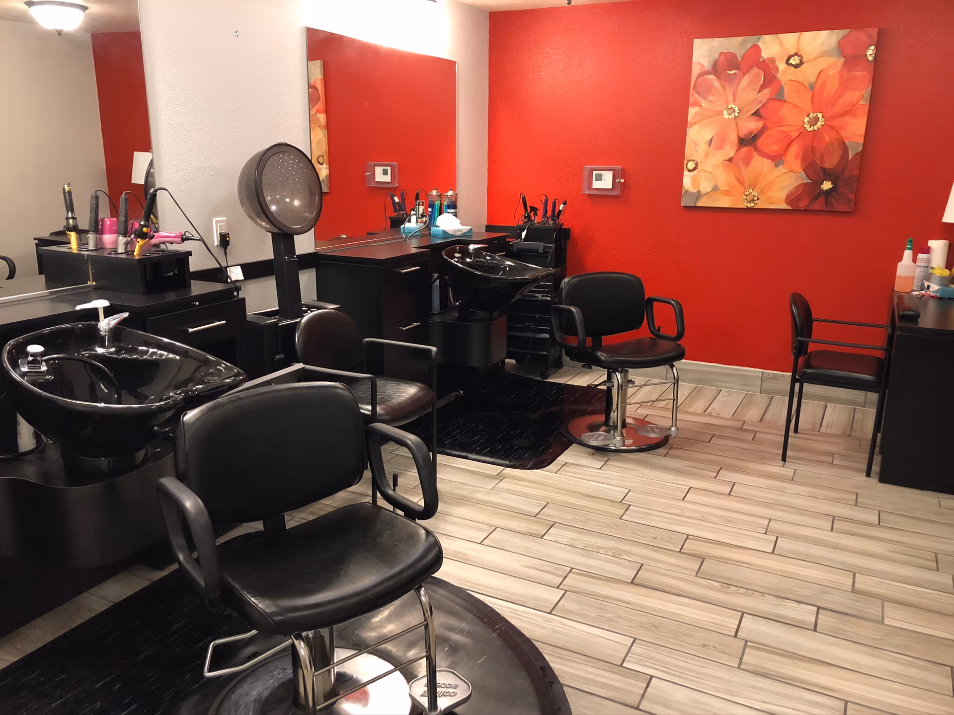 Interior of a salon area with black salon chairs, hair washing sinks, hair dryers, and a large mirror on the wall. The walls are painted red and white, with a floral painting hanging on the red wall. The floor is tiled with light wood-like tiles.