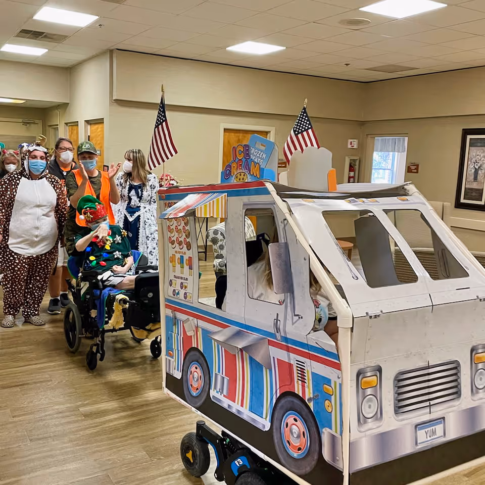 A group of people in costumes and masks are gathered indoors around a wheelchair decorated as an ice cream truck with American flags on top. The setting appears to be a senior living facility with beige walls and wooden floors.