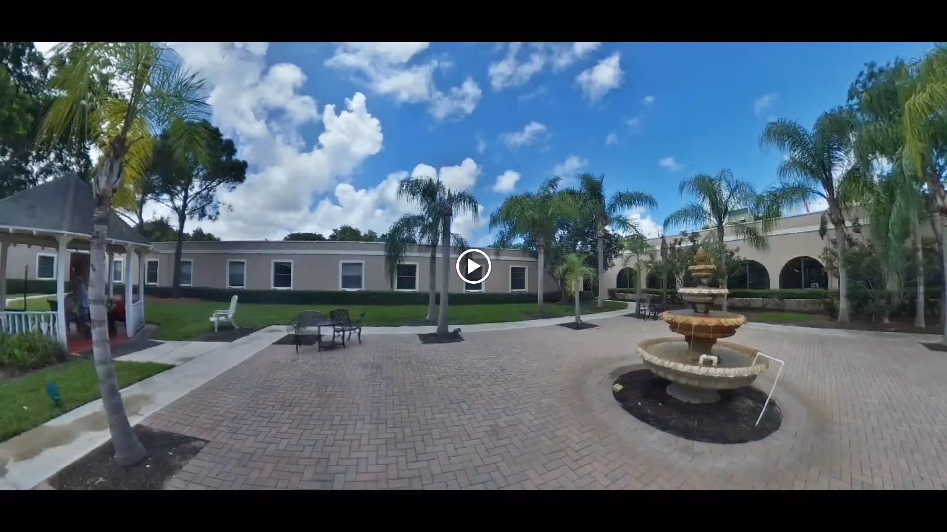 Courtyard with a central tiered fountain, palm trees, a gazebo and seating in front of a single-story building under a blue sky.
