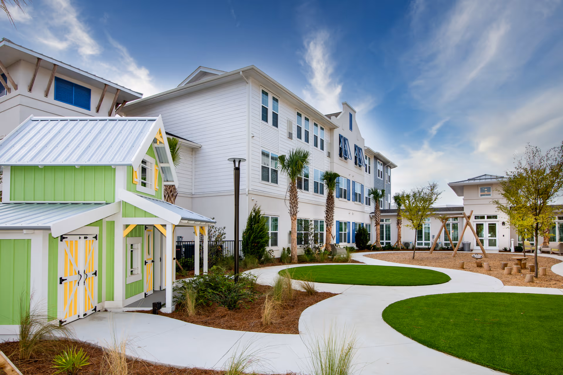 Exterior view of a white multi-story senior living building and courtyard with palm trees, circular lawns, walkways, and a small green playhouse.