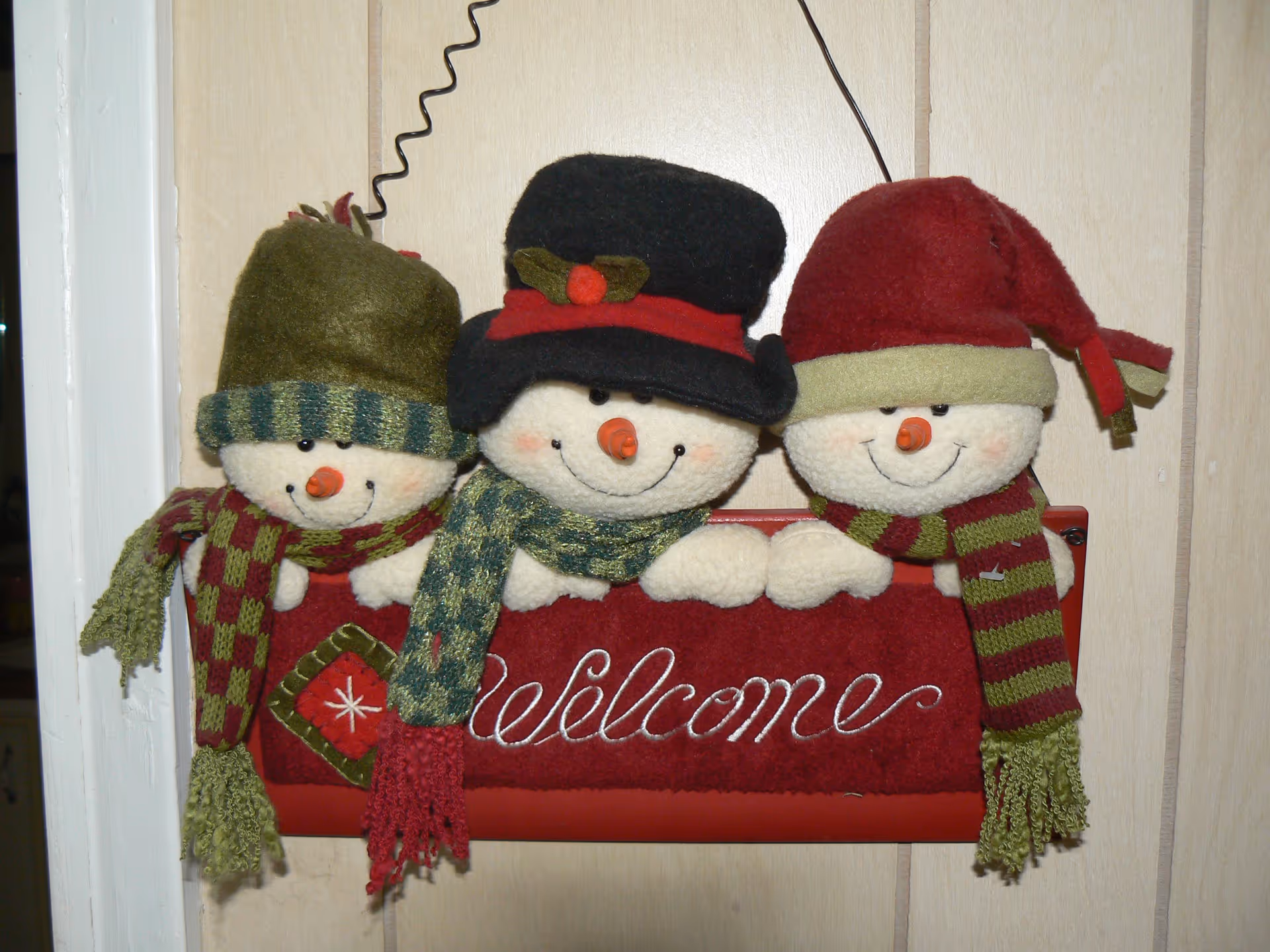 Three plush snowmen wearing hats and scarves perched above a red 'Welcome' sign on a wood-paneled wall.