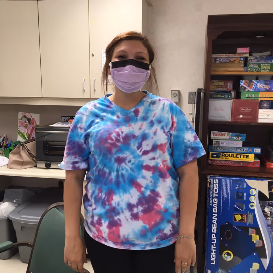 A woman wearing a purple and black face mask and a blue, purple, and white tie-dye shirt stands in a room with cabinets, a toaster oven, and a shelf filled with board games and other items.