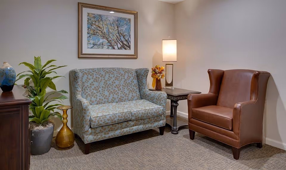 A cozy seating area with a patterned loveseat and a brown leather armchair. Between them is a small wooden side table with a modern lamp and a small floral arrangement. On the left side, there is a wooden cabinet with a blue vase and a green potted plant. A framed painting of tree branches with blue sky hangs on the wall above the loveseat.