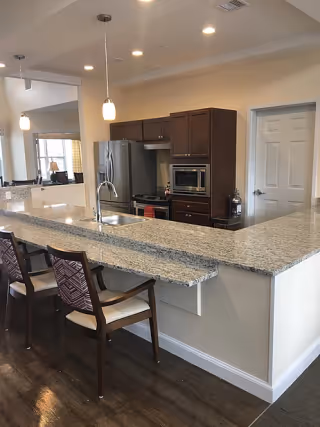 Modern open kitchen with a long granite breakfast bar, two upholstered barstools, stainless steel refrigerator, and built-in microwave.