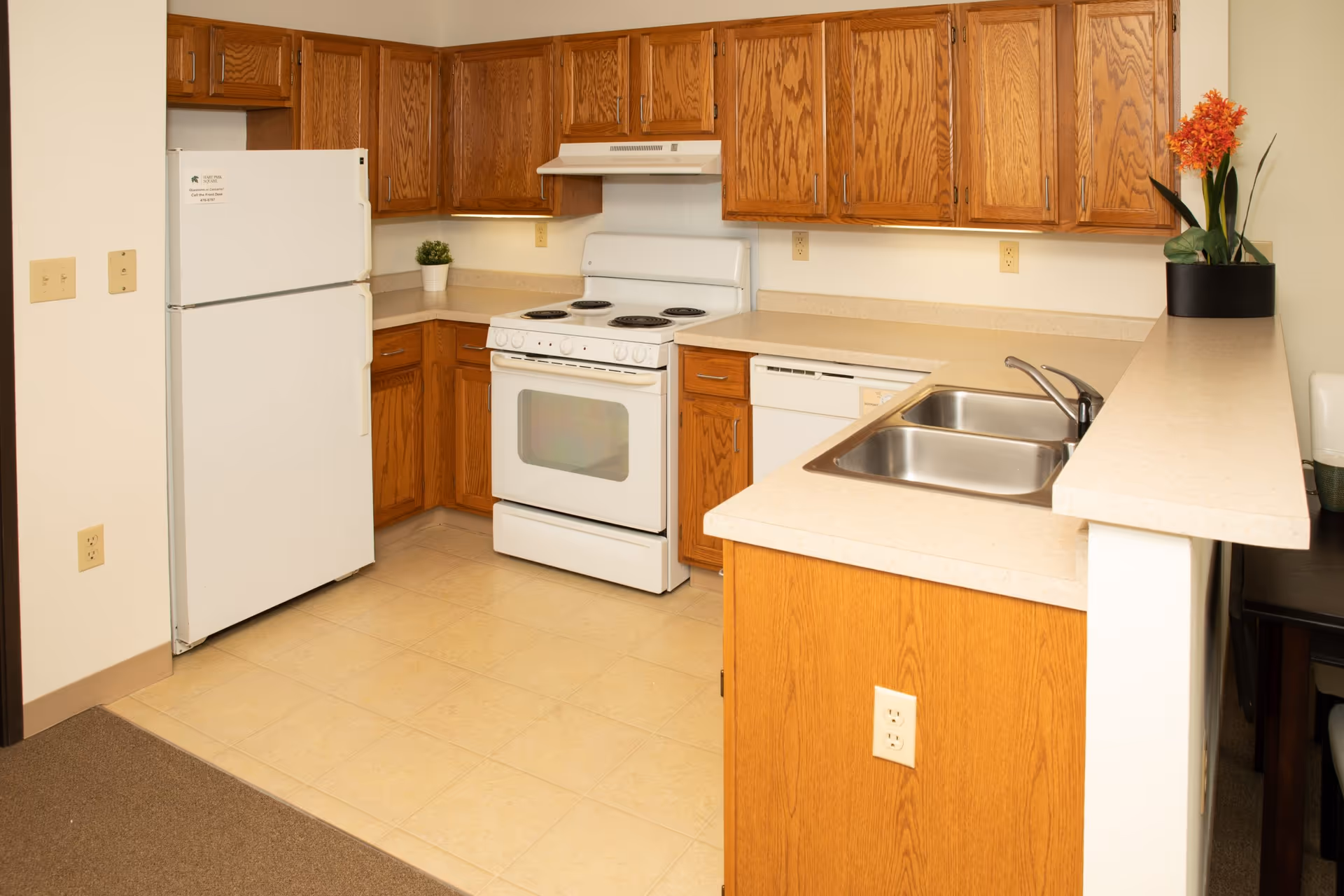 A kitchen with wooden cabinets, a white refrigerator, a white electric stove with an oven, a white dishwasher, and a double stainless steel sink. There is a small potted plant on the countertop near the refrigerator and a black pot with orange flowers on the counter near the sink.