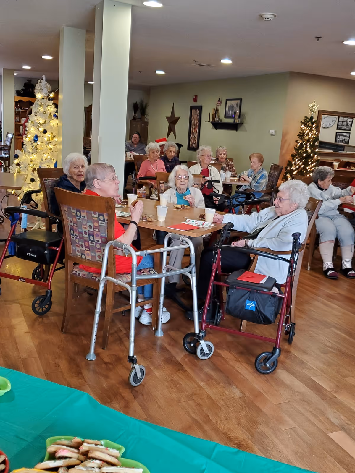 A group of elderly women sitting around tables in a communal dining area decorated with Christmas trees and holiday decorations. Some women have walkers nearby, and there are plates and cups on the tables. The room has wooden flooring and soft lighting.