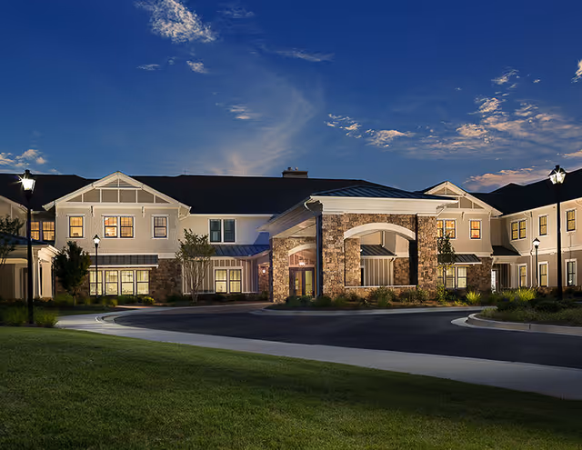 Exterior view of The Glen at Lake Oconee Village senior living facility at dusk, showing a large two-story building with stone and white siding, illuminated windows, street lamps, and a circular driveway surrounded by green grass.