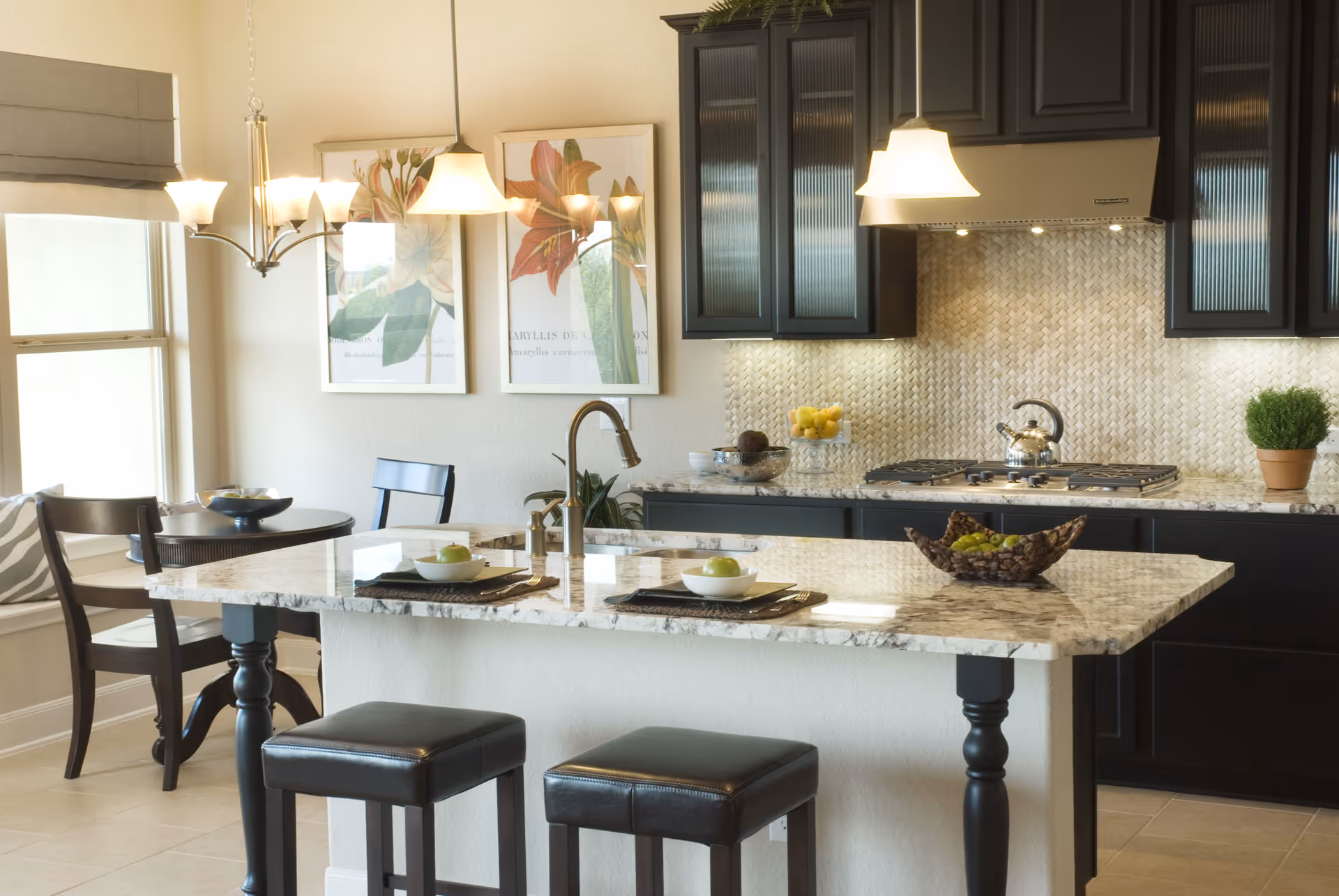 A modern kitchen with a marble countertop island featuring two black leather stools. The kitchen has dark cabinetry, a stainless steel stove with a kettle, a decorative backsplash, and pendant lights hanging above the island. There is a small round dining table with two chairs near a window, and framed floral artwork on the wall.