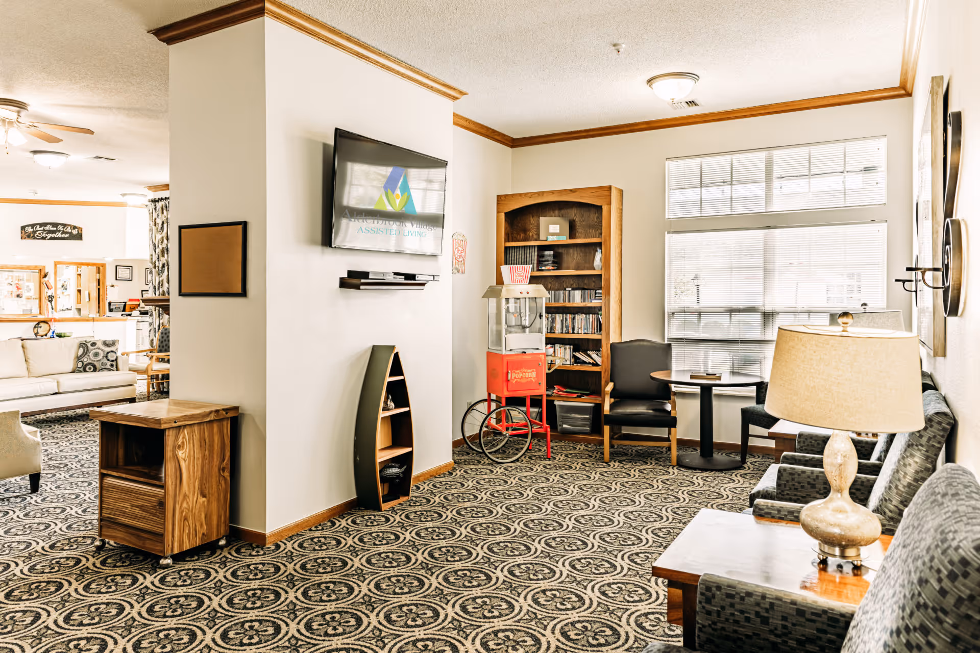 A bright and cozy living room area in Alderbrook Village assisted living facility featuring patterned carpet, multiple seating options including armchairs and sofas, a wooden bookshelf filled with DVDs, a vintage-style popcorn machine, a small round table with chairs near a large window with blinds, a wall-mounted TV displaying the Alderbrook Village logo, and a table lamp on a side table.