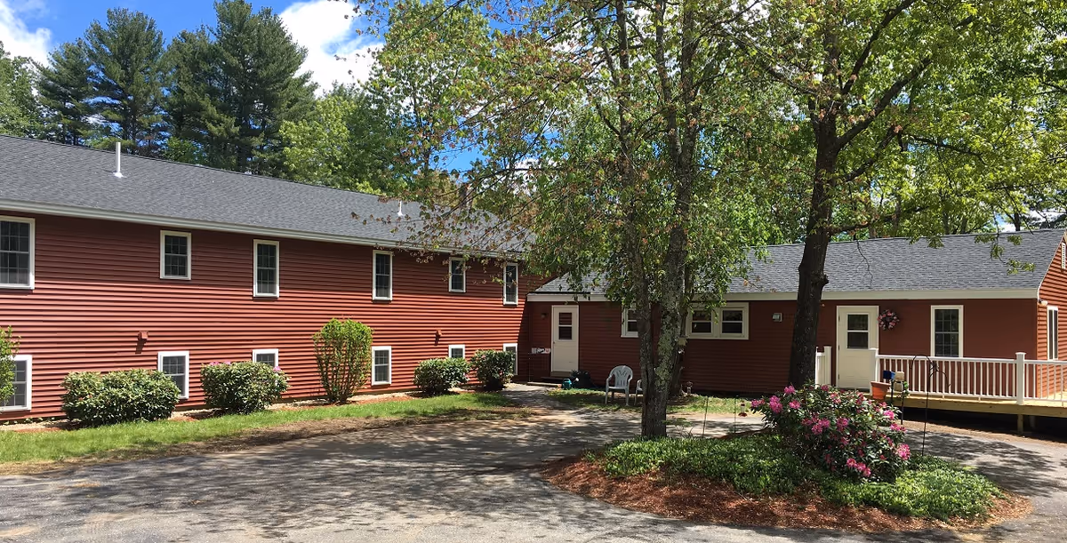 Exterior view of a red two-story building with white trim, surrounded by green trees and bushes under a blue sky with some clouds. There is a paved driveway and a small garden area with flowers and mulch in front of the building.