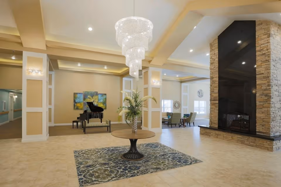 Spacious senior living facility common area with a large chandelier hanging from the ceiling, a round table with a floral arrangement on a patterned rug, a grand piano with a bench against the wall, and a stone fireplace with a black mantle. There are seating areas with chairs and tables near windows in the background.