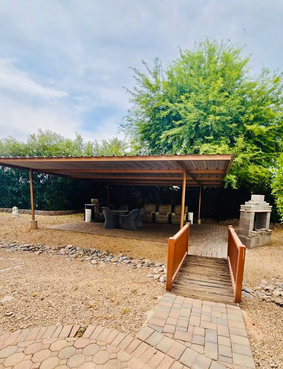 Outdoor seating area with a wooden bridge leading to a covered patio that has chairs and a table. There is a stone fireplace on the right side and trees and shrubs surrounding the area under a partly cloudy sky.