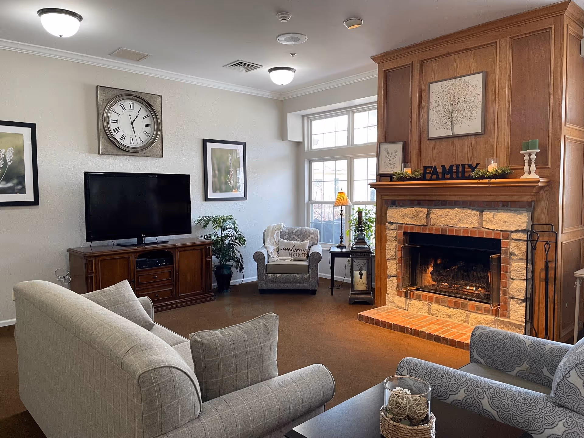 A cozy living room with a lit stone and brick fireplace on the right, decorated with framed pictures, candles, and a 'FAMILY' sign on the mantel. There is a large window behind a gray armchair with a 'welcome HOME' pillow, a wooden TV stand with a flat-screen TV, a wall clock above it, and two framed pictures on the wall. The room features a beige sofa and a patterned armchair with a coffee table holding a decorative glass container with woven balls.