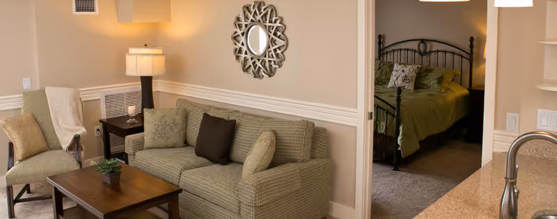 A cozy living room with a patterned sofa, two cushions, a wooden coffee table with a small plant, and a chair with a throw blanket. A decorative round mirror hangs on the wall above the sofa. Through an open doorway, a bedroom with a bed covered in green bedding is visible. The edge of a kitchen counter with a faucet is seen on the right side.