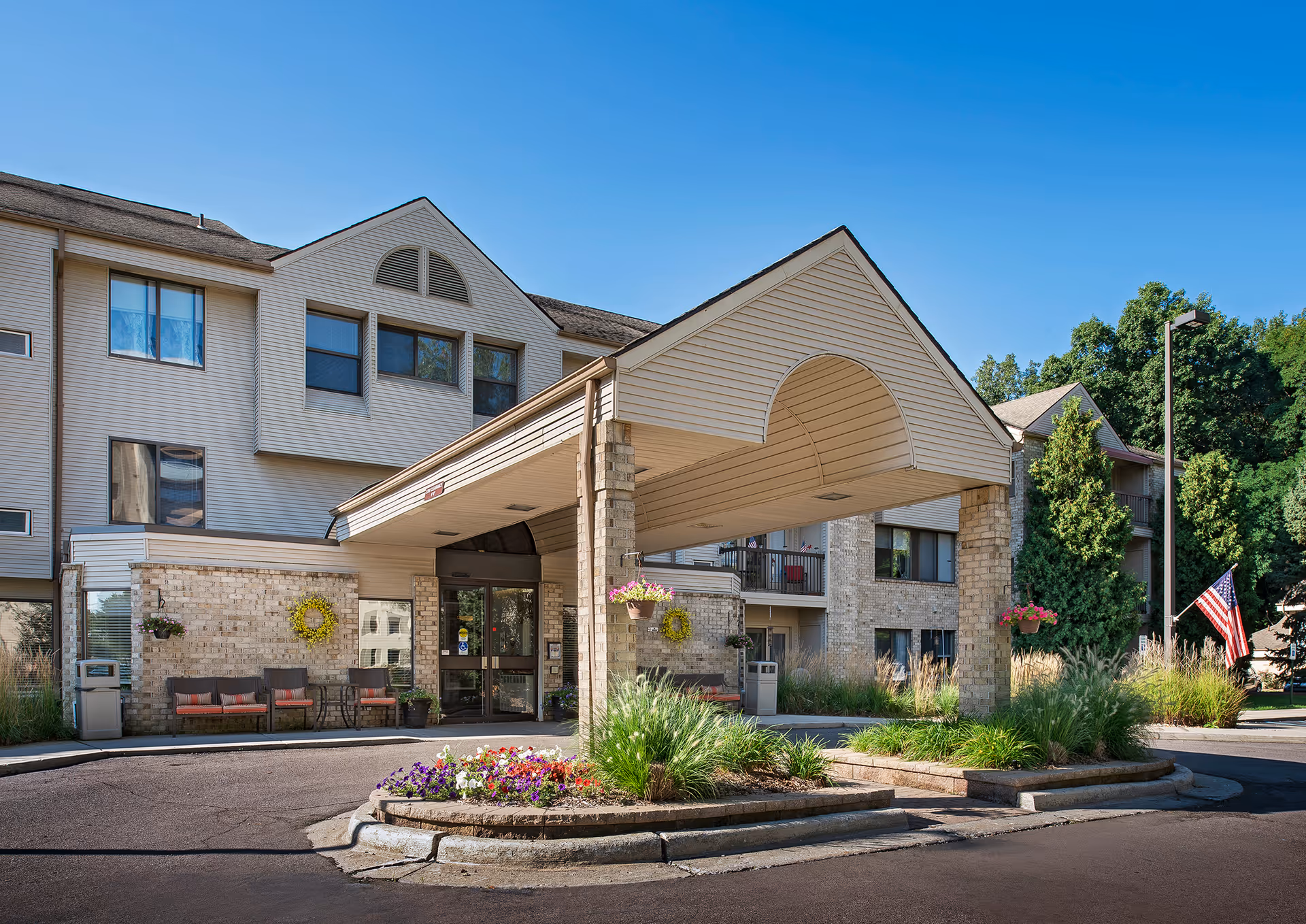 Exterior view of a senior living facility named American House Carpenter with a covered entrance, brick and siding facade, outdoor seating, flower beds, and an American flag on a sunny day.