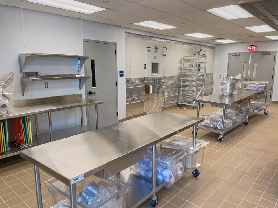 A clean, industrial kitchen or food preparation area with stainless steel tables on wheels, metal shelving, and storage bins containing various kitchen supplies. The room has tiled floors, white walls, and fluorescent ceiling lights. Double doors and a small windowed door are visible at the far end.
