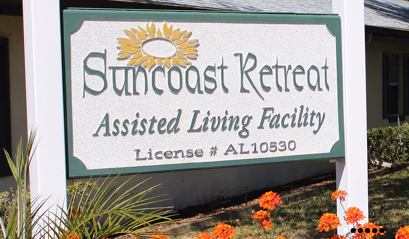 A white and green sign for Suncoast Retreat Assisted Living Facility with a sunflower graphic above the name, surrounded by some plants and orange flowers in front of a building.