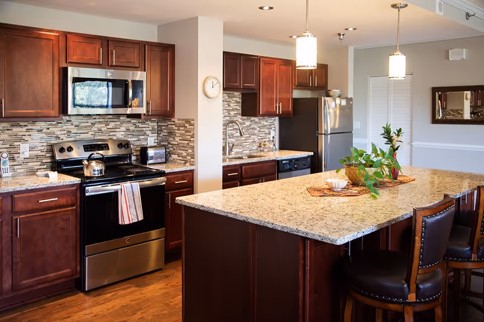 A modern kitchen with dark wooden cabinets, stainless steel appliances including a microwave, stove, and refrigerator. The kitchen features a large granite island with three high-back chairs, pendant lighting, and decorative plants on the island. The backsplash has a mosaic tile design, and the floor is wooden.