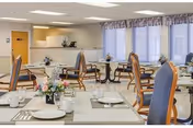 Bright dining room with multiple set tables and upholstered chairs, floral centerpieces, large windows and a serving counter in the back.