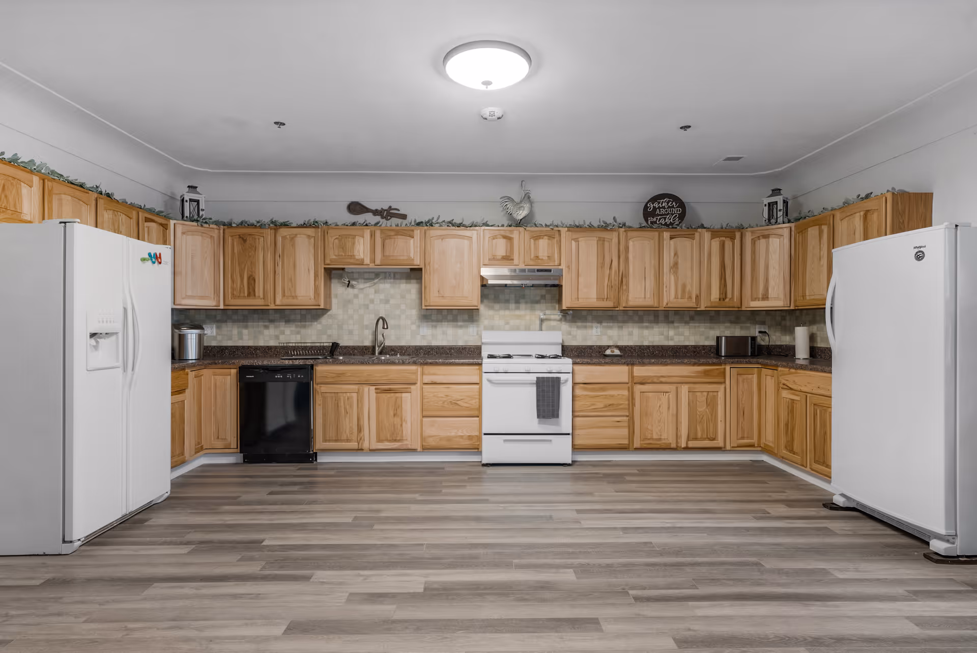 A spacious kitchen with wooden cabinets lining the walls, two white refrigerators on either side, a white stove with a gray towel hanging on the handle, a black dishwasher, and a sink in the middle. The kitchen has a light wood floor and a ceiling light fixture. Decorative greenery and kitchen-themed decor are placed above the cabinets.