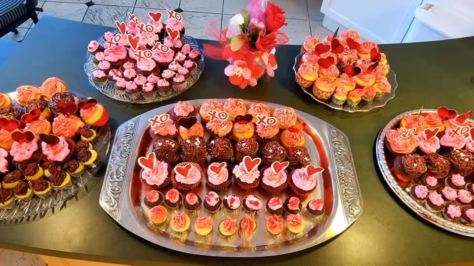 A variety of decorated cupcakes arranged on multiple trays on a kitchen counter. The cupcakes have pink, red, and chocolate frosting with heart-shaped and XO decorations. A small vase with red and white flowers is placed among the trays.