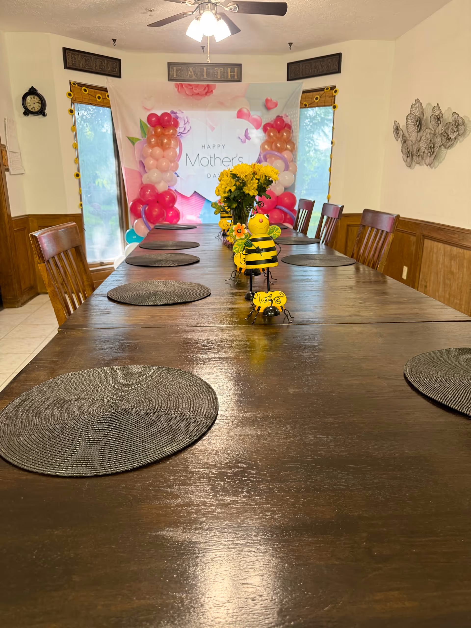 A long wooden dining table with round black placemats and decorative yellow bee figurines in the center. A vase with yellow flowers is also on the table. In the background, there is a Mother's Day banner with pink and red balloons on either side. The room has wooden chairs, two windows with yellow curtains, a ceiling fan with lights, and wall decorations including a clock and metal floral art.