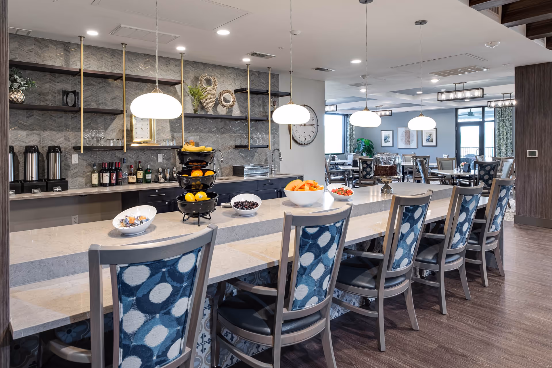 Bright communal dining area with a long marble counter, patterned bar stools, pendant lights, and bowls of fruit and snacks.