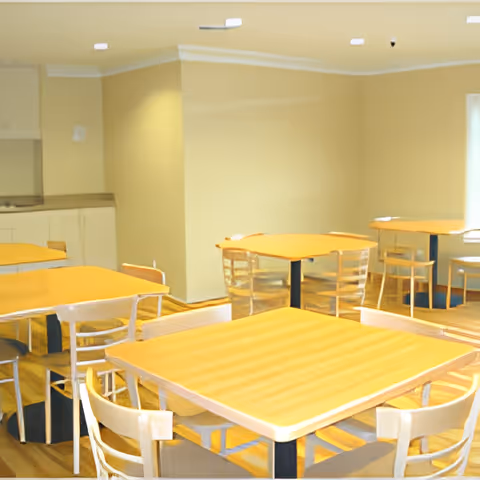A bright dining area with multiple wooden tables and chairs arranged neatly. The room has light-colored walls and a wooden floor. There is a counter with cabinets along one wall and ceiling lights providing illumination.
