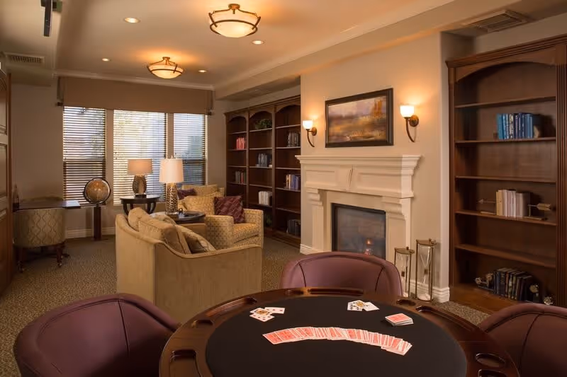 A cozy living room area with a round poker table in the foreground featuring playing cards, surrounded by four purple chairs. In the background, there is a beige sofa and armchair with patterned cushions, a fireplace with a painting above it, two wall sconces, and built-in wooden bookshelves filled with books. Large windows with blinds and two table lamps provide natural and artificial light.