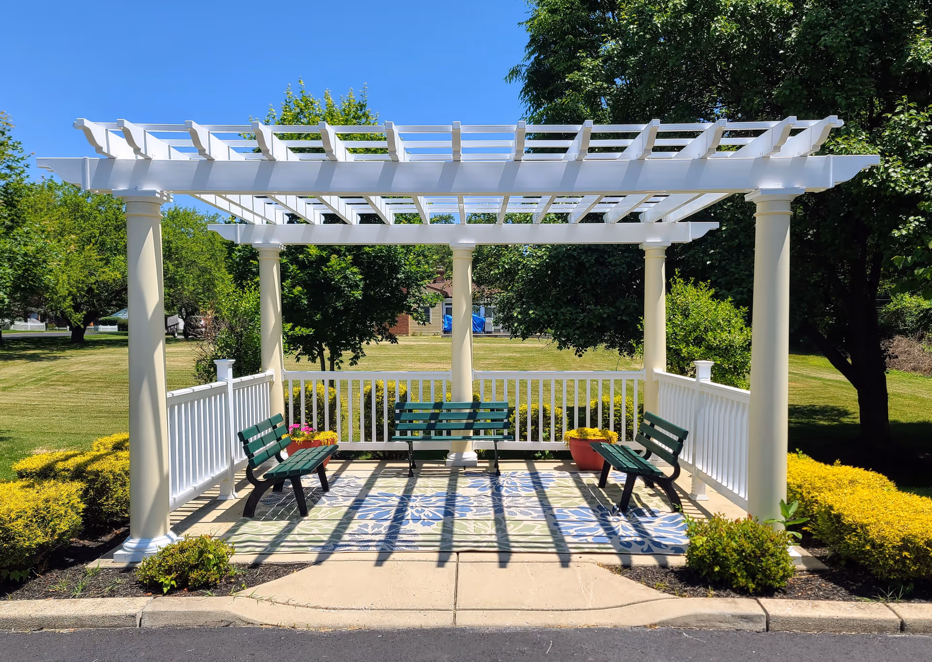 White pergola with green benches, potted flowers and a patterned rug set on a paved pad in a sunny grassy garden.