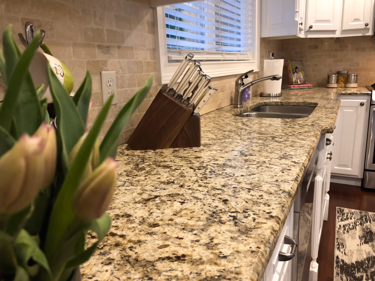 A kitchen countertop with a granite surface, a knife block with knives, a stainless steel sink with a faucet, a paper towel holder, and white cabinets. There is a window with blinds above the sink and a blurred bouquet of flowers in the foreground on the left side.