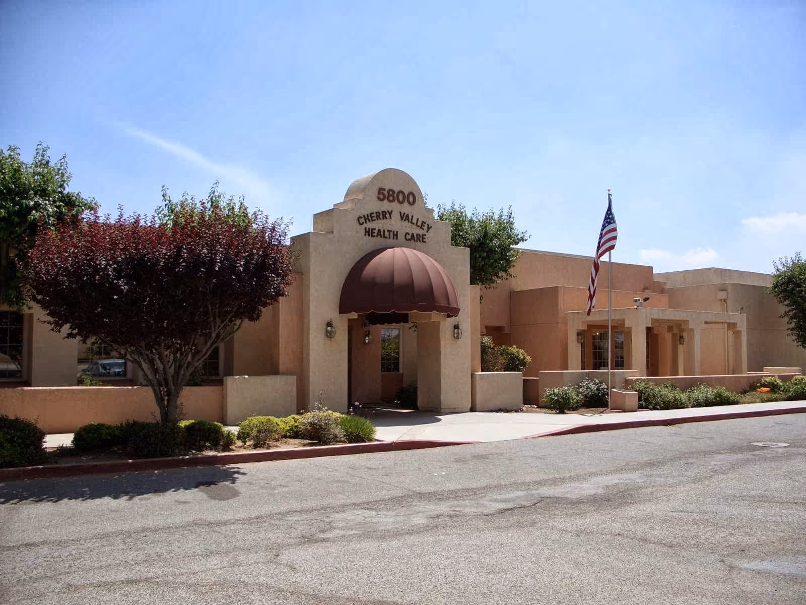 Exterior front view of Cherry Valley Health Care building with beige stucco walls, a brown awning over the entrance, an American flag on a flagpole, and landscaping including trees and bushes under a clear blue sky.