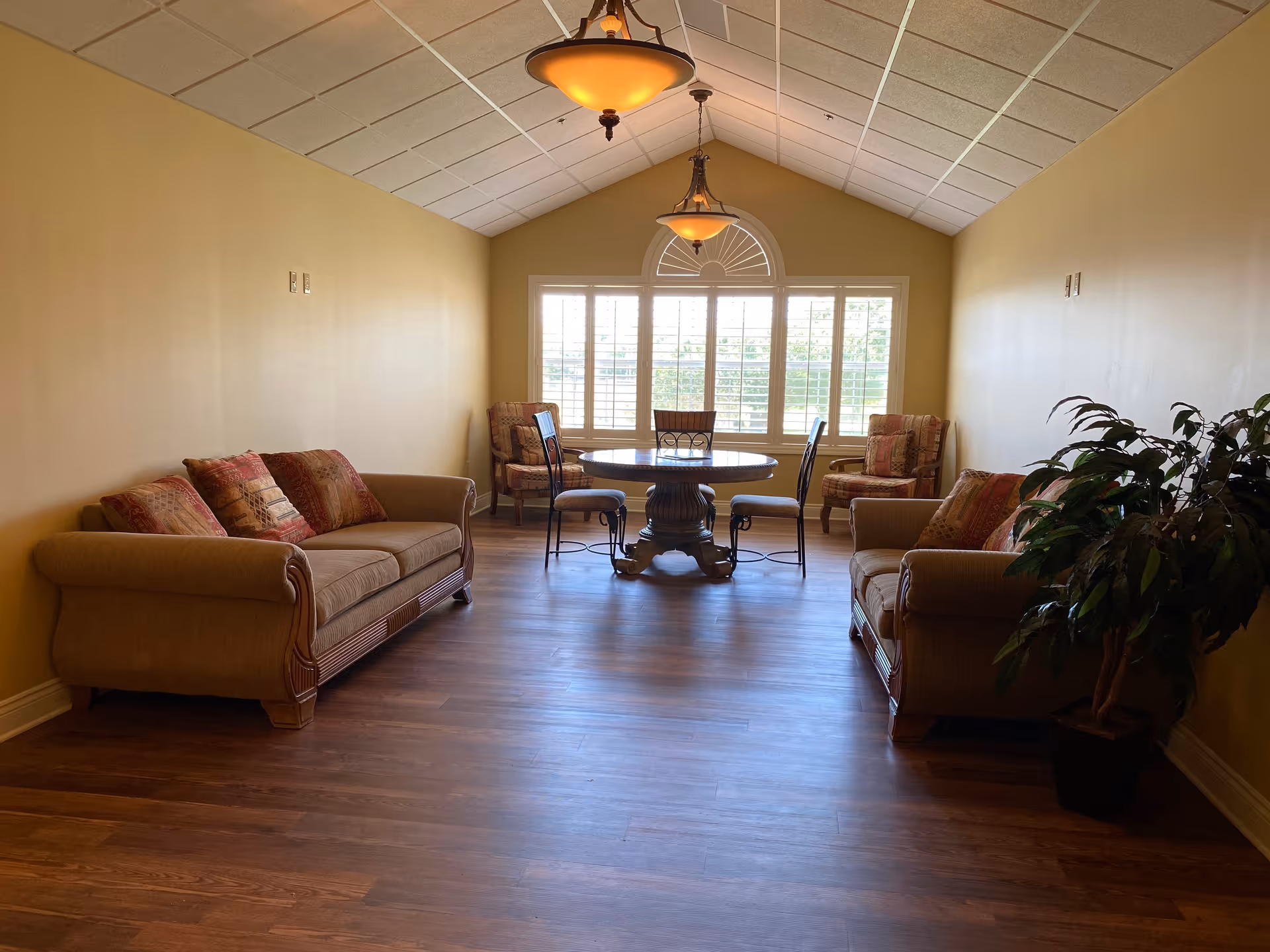 A cozy living room with two beige sofas adorned with patterned cushions facing each other, a round wooden table with four chairs near a large window with white shutters, warm yellow walls, wooden flooring, two hanging ceiling lights, and a potted plant in the corner.
