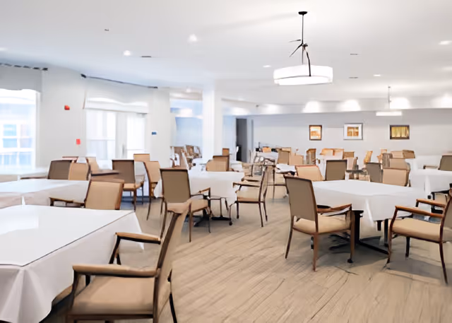 Bright communal dining room with multiple tables covered in white linens and wooden chairs.