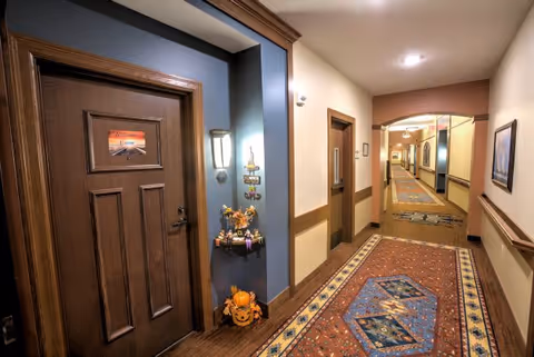 A well-lit hallway in a senior living facility with wooden doors and trim. The hallway features a patterned carpet runner and light-colored walls with wooden chair rails. One door has a small shelf next to it decorated with autumn and Halloween-themed items, including a pumpkin and a small scarecrow. The hallway extends into the distance with more doors and framed artwork on the walls.