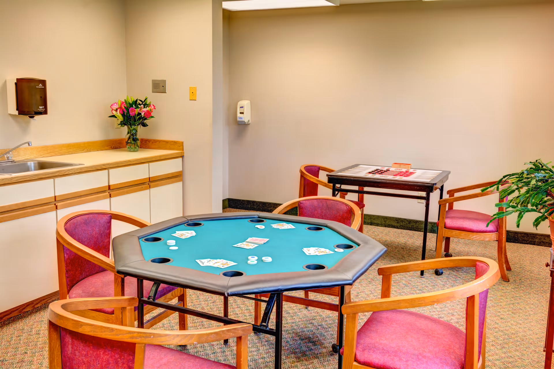 A game room with a poker table set up with playing cards and poker chips, surrounded by four wooden chairs with red cushions. In the background, there is a smaller table with a checkerboard and two chairs, a countertop with a sink, a vase of flowers, a wall-mounted soap dispenser, and a potted plant on the right side.