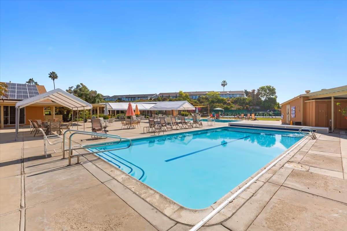 Outdoor swimming pool with lounge chairs, shaded seating areas, and a concrete patio under a clear blue sky.