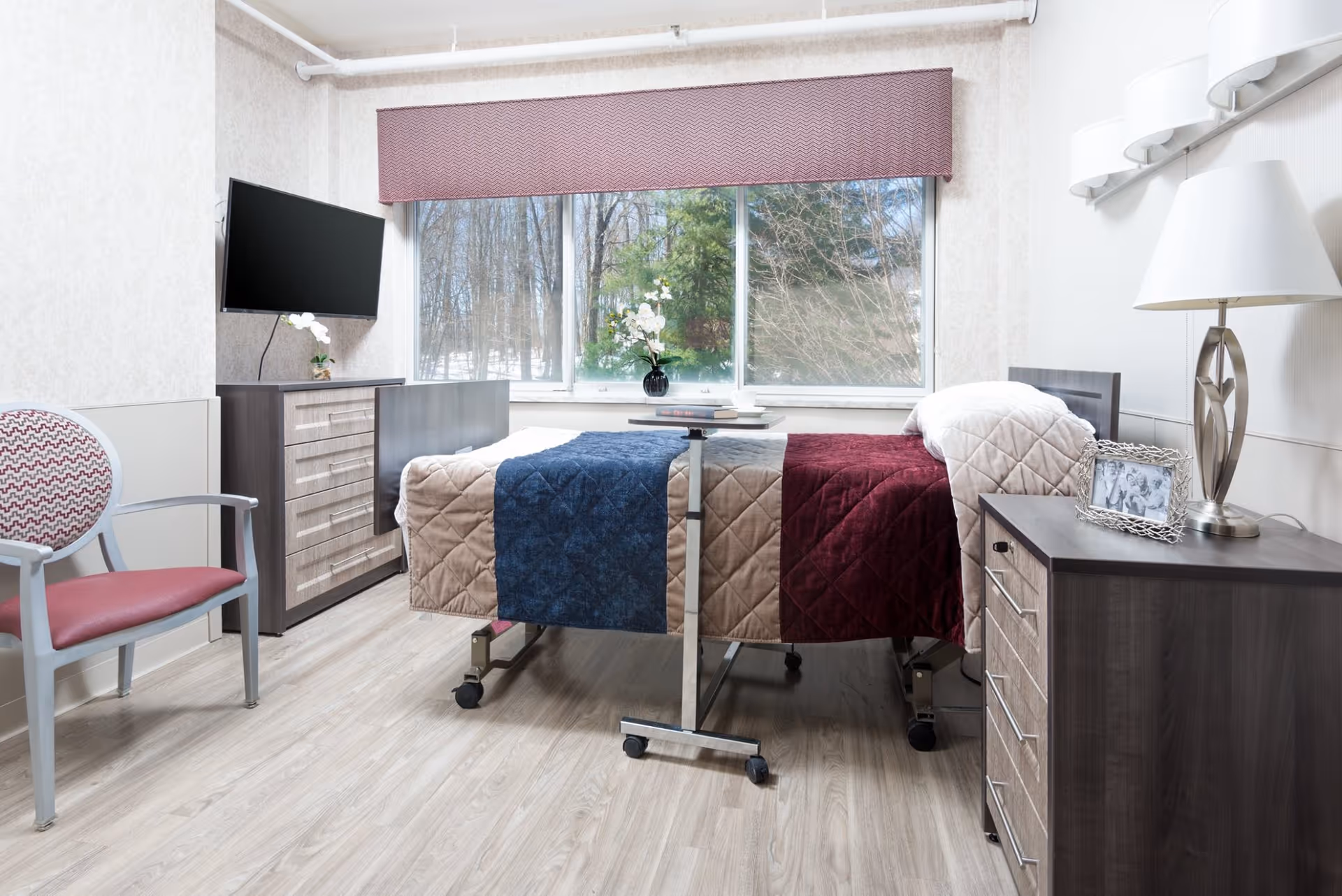 A bright senior living bedroom featuring a hospital-style bed with quilted blankets, bedside tables, a wall-mounted TV, a chair, and a large window overlooking trees.
