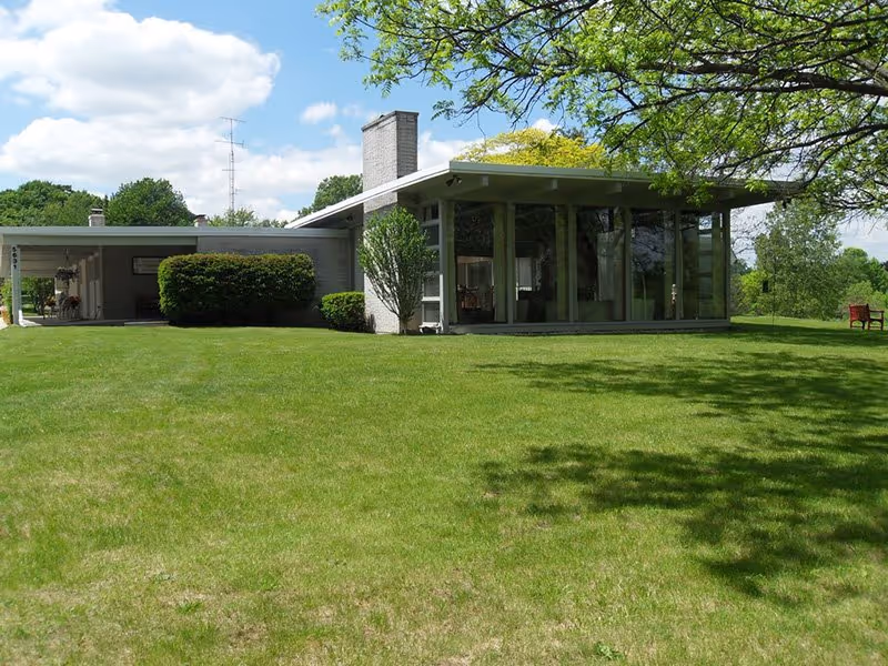 A modern single-story building with large glass windows and a flat roof, surrounded by a well-maintained green lawn and trees under a partly cloudy blue sky.