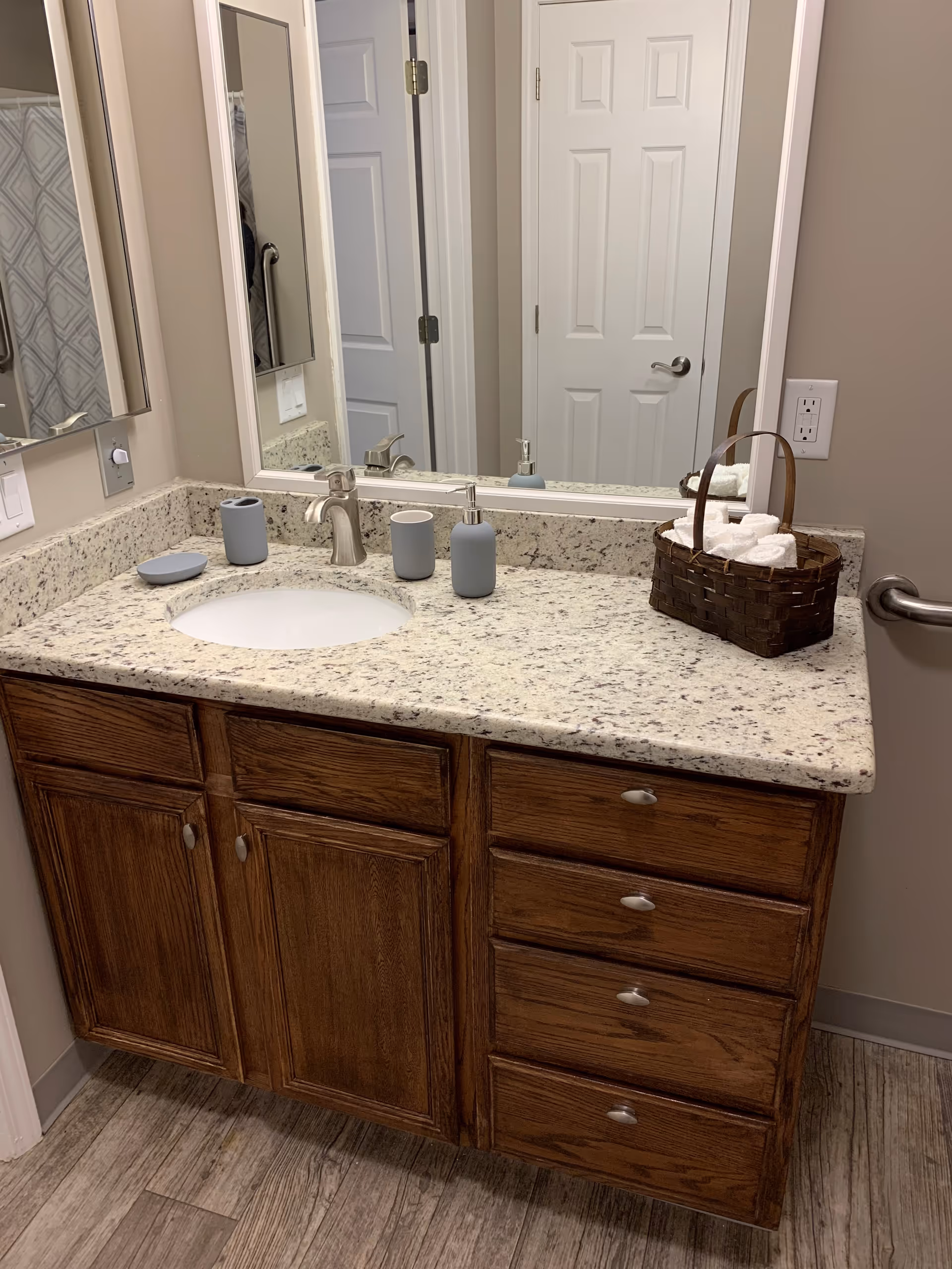 A bathroom vanity with a granite countertop, an under-mount sink, and a silver faucet. On the countertop are a soap dish, a toothbrush holder, a soap dispenser, and a basket filled with rolled white towels. A large mirror is mounted on the wall above the vanity, reflecting a closed white door and part of a shower curtain with a geometric pattern. The vanity has wooden cabinets and drawers with silver handles, and the floor has wood-like tiles.
