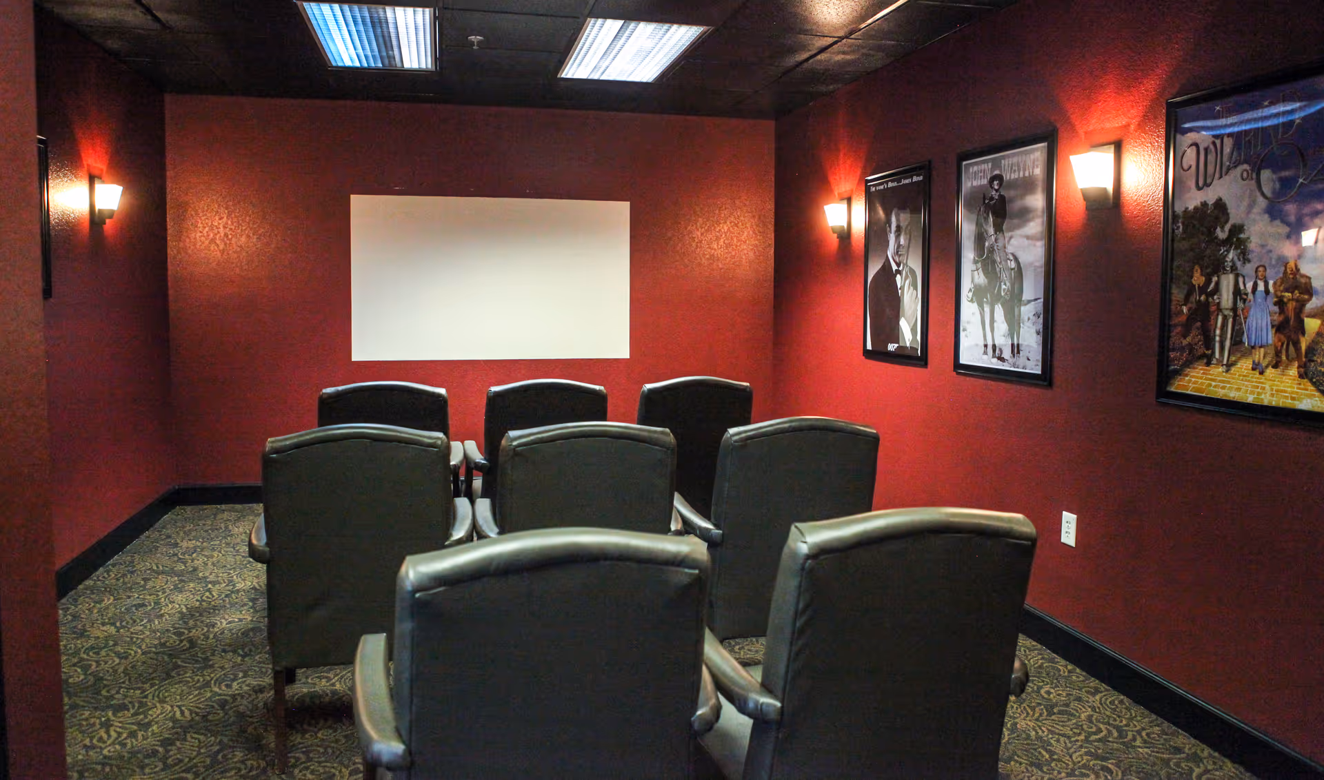 A small screening room with two rows of leather recliner chairs facing a blank projection screen, red walls, and framed movie posters.