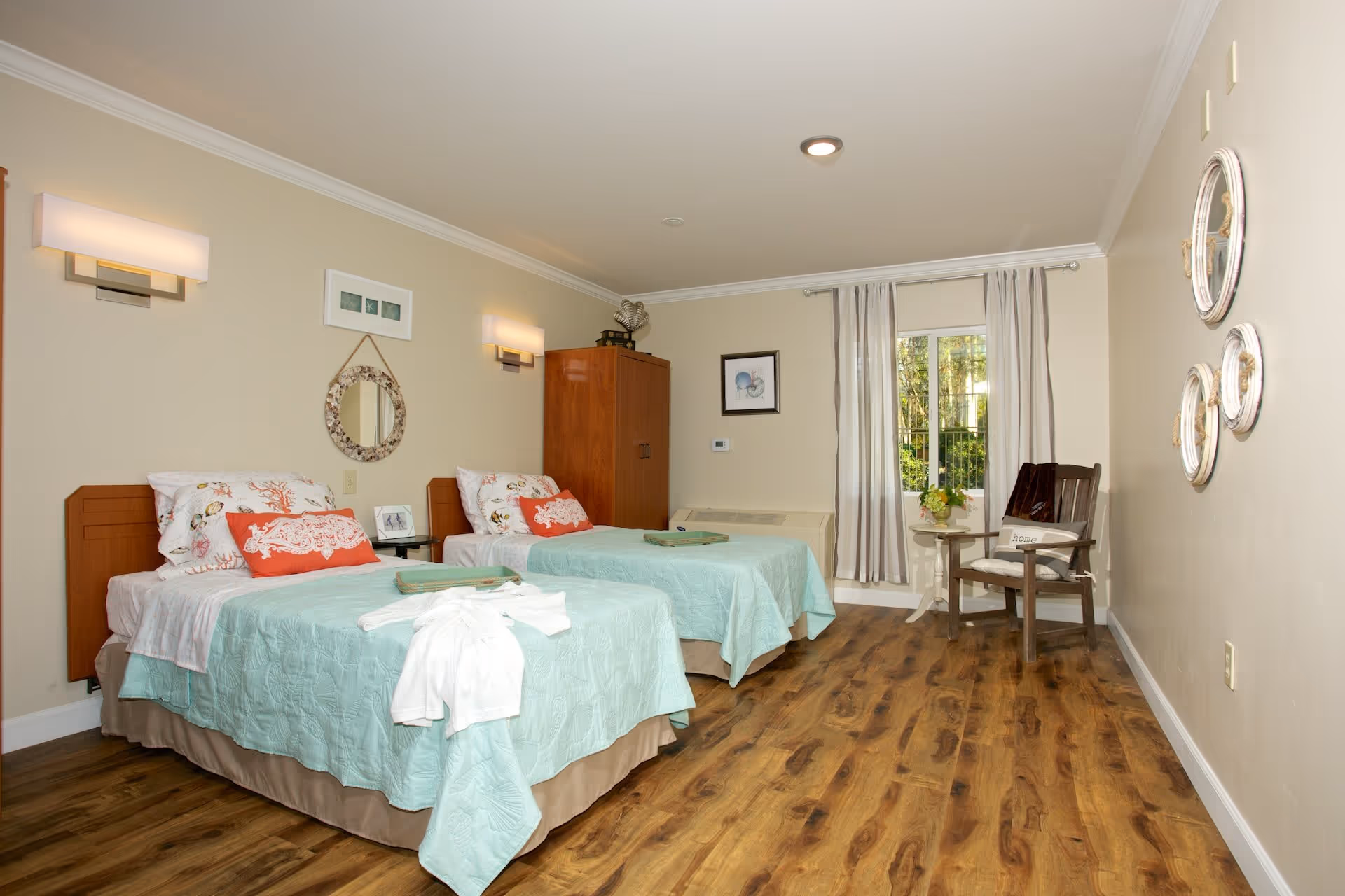 A bright and clean bedroom with two twin beds, each made with light blue bedspreads and decorative pillows. The room features wooden flooring, a wooden cabinet, a small side table with a flower vase, a wooden chair with a cushion, and a window with light curtains letting in natural light. The walls are decorated with a round mirror and framed pictures.