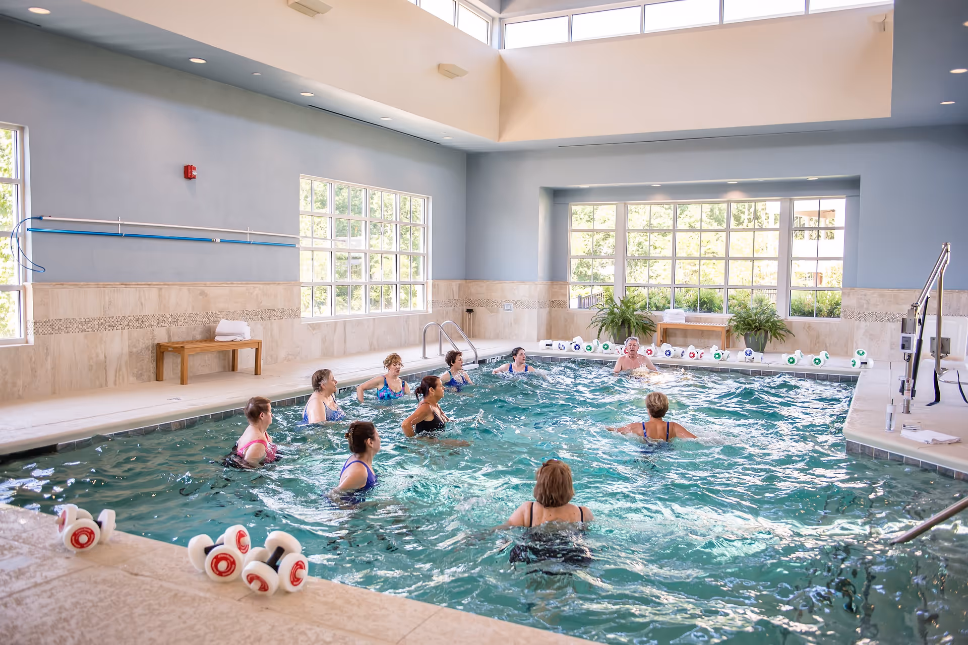 A group of elderly people participating in a water exercise class in an indoor swimming pool with large windows letting in natural light. The pool area has light blue walls, beige tiled floors, and aquatic exercise equipment on the pool deck.