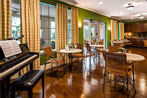 A cozy interior common area with wooden floors, green walls, and large windows covered by patterned curtains. There are several round tables with wicker chairs arranged around them, each table decorated with a small flower arrangement. A black piano with sheet music is positioned on the left side of the room. In the background, there is a kitchen area with dark wooden cabinets and warm lighting.