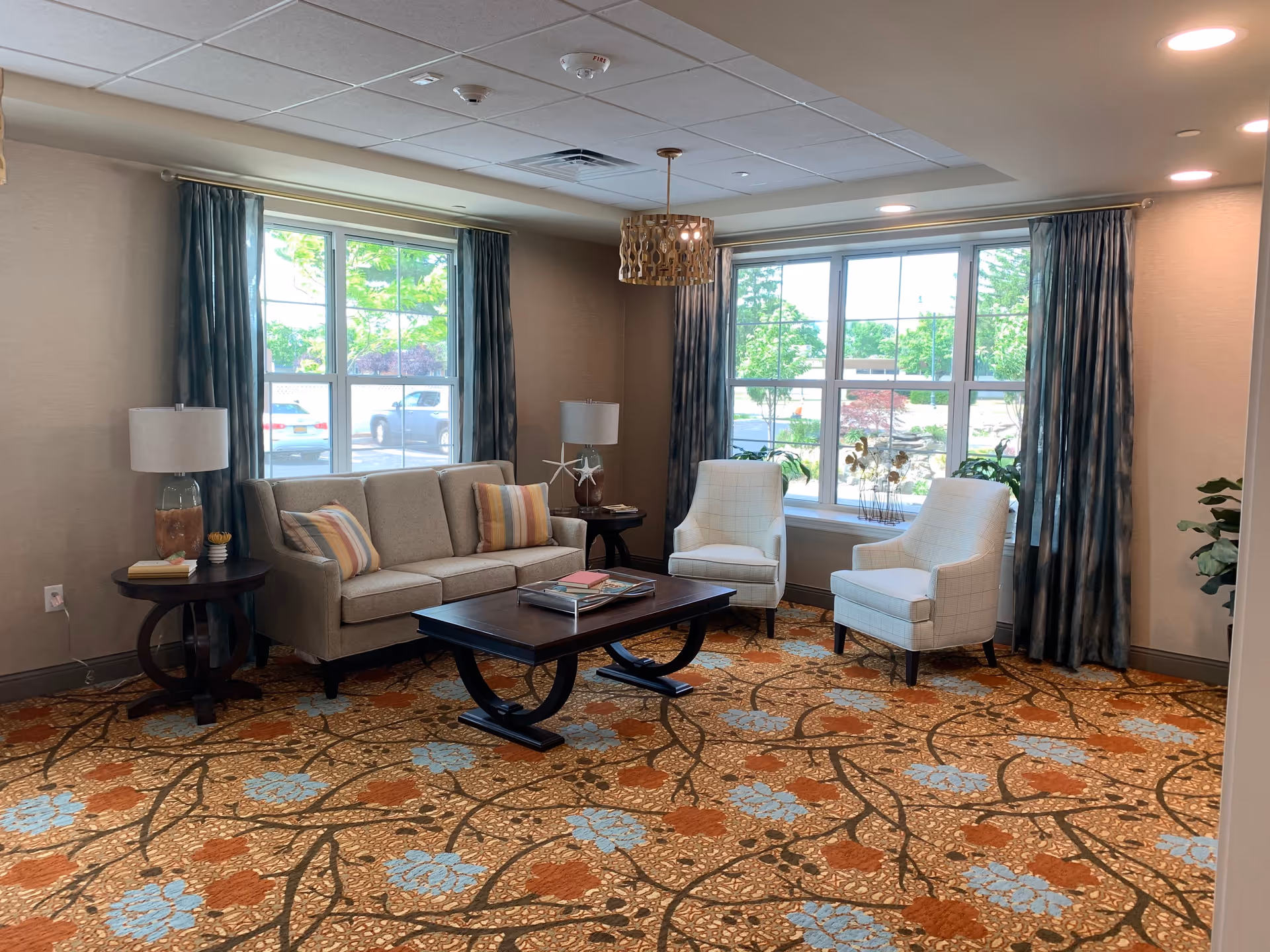 A cozy living room area with a beige sofa adorned with striped pillows, two white armchairs, a dark wooden coffee table with books on it, and two side tables each with a lamp. The room has large windows with blue curtains letting in natural light, and a patterned carpet with orange and blue floral designs.